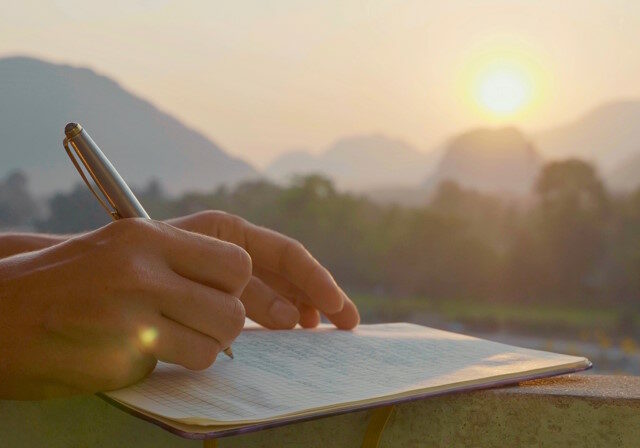 Young woman writing travel notes in diary during sunrise with beautiful sun light and mountain landscape on the background, close-up.